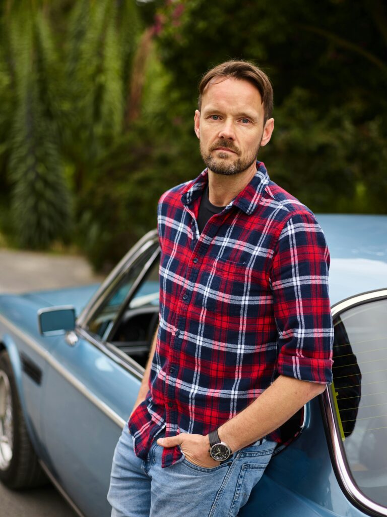 Adult pensive bearded guy in wristwatch standing with hand on pocket leaned on blue retro car near green trees in countryside and looking at camera
